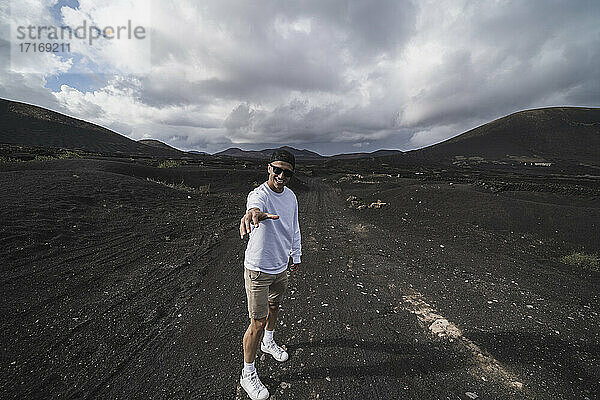 Lächelnder männlicher Tourist  der auf dem Vulkan El Cuervo  Lanzarote  Spanien  auf schwarzer Erde steht und gestikuliert