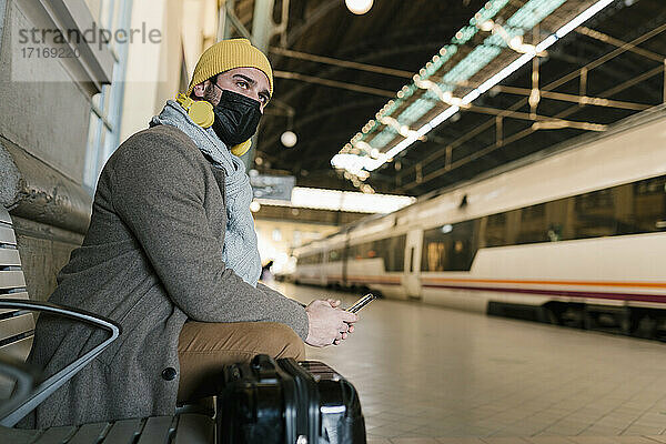 Mann mit Gesichtsschutz  der auf einer Bank im Bahnhof sitzt und ein Mobiltelefon benutzt