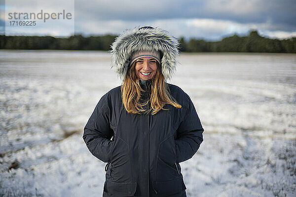 Lächelnde schöne Frau mit Händen in den Taschen  die auf einem verschneiten Feld vor dem Himmel steht