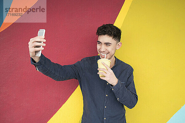 Junger Mann  der ein Selfie mit seinem Smartphone macht  während er Saft gegen eine bunte Wand trinkt