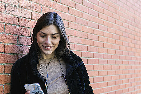 Smiling woman with mobile phone listening music through headphones while leaning brick wall