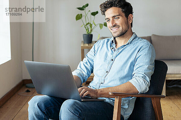 Lächelnder Mann  der einen Laptop benutzt  während er zu Hause auf einem Sessel sitzt