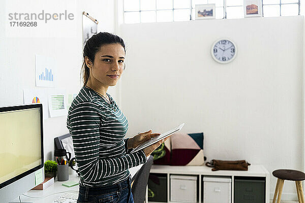 Lächelnde kreative Geschäftsfrau  die ein digitales Tablet hält  während sie im Büro steht