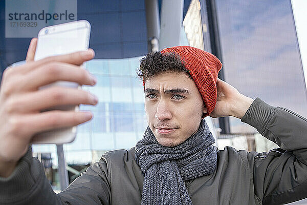Junger Mann  der sein Strickzeug anpasst  während er ein Selfie mit seinem Smartphone in der Stadt macht