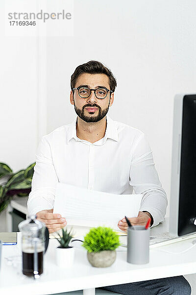 Männlicher Unternehmer  der ein Papierdokument hält  während er auf einem Schreibtisch im Büro sitzt