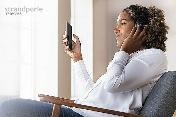 Mittlere erwachsene Frau mit Mobiltelefon hört Musik über Kopfhörer  während sie zu Hause auf einem Sessel sitzt