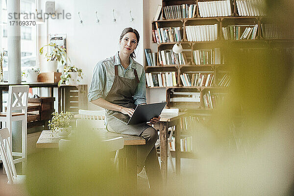 Weiblicher Manager in Schürze mit Laptop  der am Tisch eines Cafés sitzt und nachdenkt