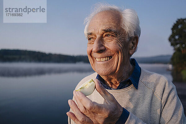 Lächelnder Mann isst Apfel  während er am See steht
