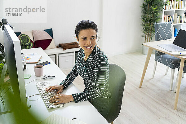 Lächelnde junge Geschäftsfrau  die wegschaut  während sie am Schreibtisch vor einem Computer im Büro sitzt