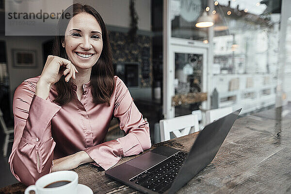 Lächelnde Unternehmerin mit Kaffeetasse und Laptop am Tisch in einem Cafe
