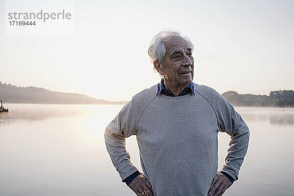 Älterer Mann mit Hand an der Hüfte  der wegschaut  während er am See steht