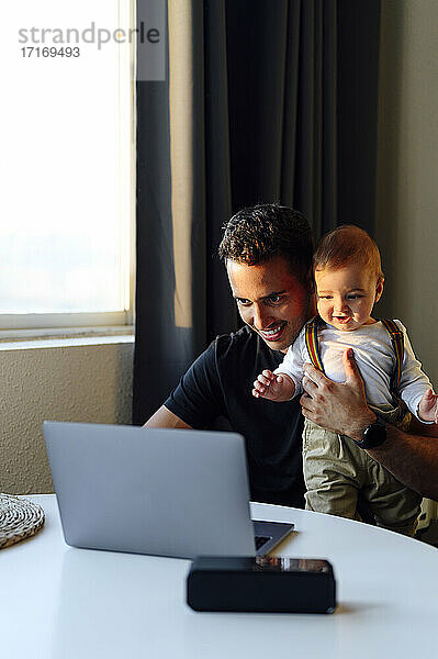 Mid adult man holding baby boy while working on laptop at home