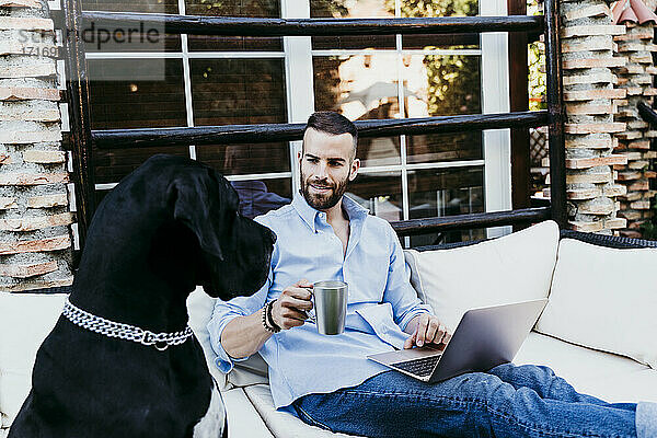 Junger Mann arbeitet am Laptop  während er mit seiner Dogge auf der Terrasse sitzt