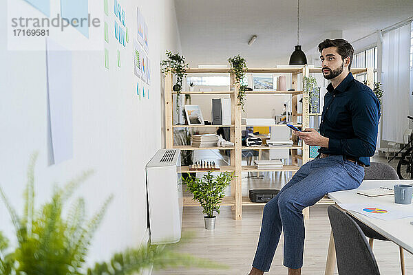 Young male entrepreneur with smart phone looking at diagram on wall while sitting on desk in office