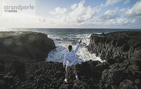 Junger männlicher Tourist  der auf einem Felsen vor dem Meer in Los Hervideros  Lanzarote  Spanien  steht und Frieden gestikuliert