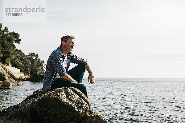 Nachdenklicher reifer Mann sitzt auf einem Felsen gegen den klaren Himmel