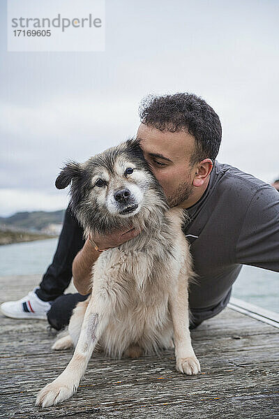 Man embracing dog while sitting on pier at beach against sky