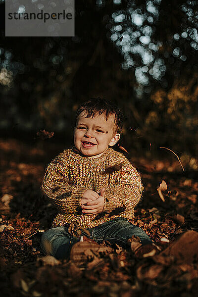 Nettes lächelndes Kleinkind sitzt inmitten trockener Herbstblätter im Wald bei Sonnenuntergang