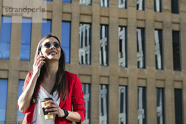 Smiling businesswoman talking on mobile phone while holding coffee cup against building