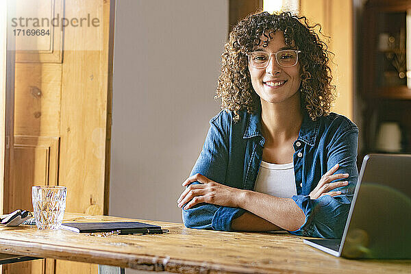 Happy young businesswoman with arms crossed by laptop at home office