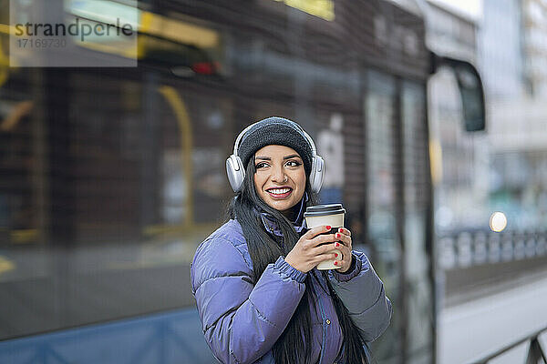 Smiling young woman with headphones holding coffee cup in city