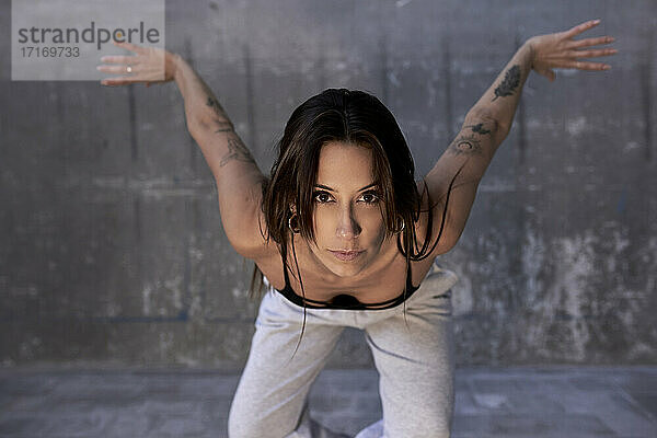 Determined female dancer staring with arms outstretched against wall in factory