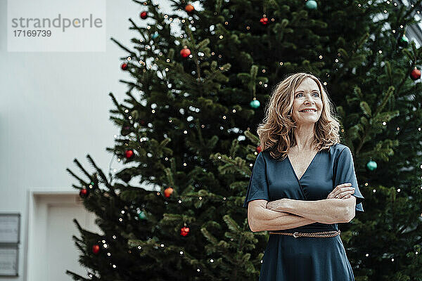 Lächelnde Geschäftsfrau mit verschränkten Armen  die gegen einen Weihnachtsbaum im Büro blickt
