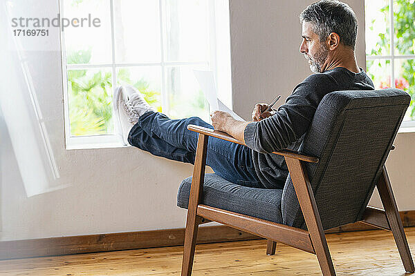 Geschäftsmann schreibt in Papier  während er mit gekreuzten Beinen auf einem Sessel zu Hause sitzt