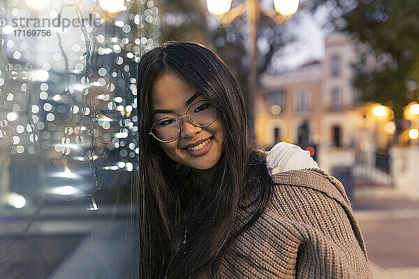 Happy young woman leaning on illuminated glass wall at dusk
