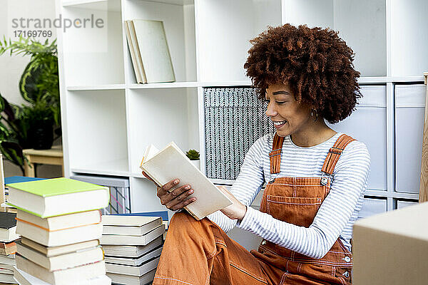 Lächelnde junge Frau  die beim Auspacken in ihrer neuen Loft-Wohnung ein Buch liest
