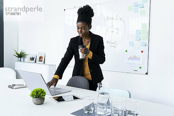 Businesswoman holding insulated drink container while using laptop at office