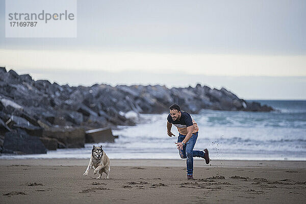 Playful man running with dog at beach against sky