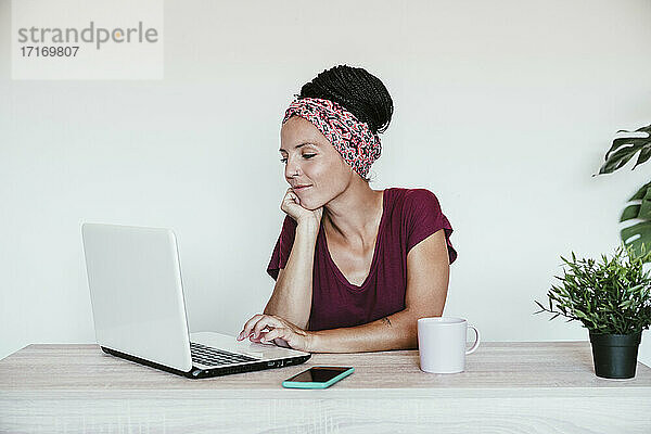 Weibliche Freiberuflerin arbeitet über einem Laptop mit der Hand auf dem Kinn gegen die Wand im Heimbüro