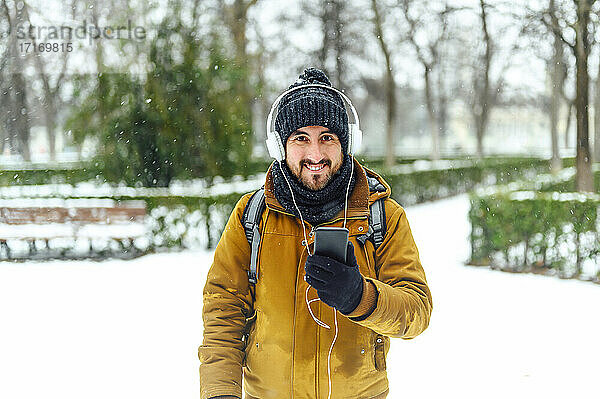 Smiling man with mobile phone listening music through headphones while standing on snow covered field during winter