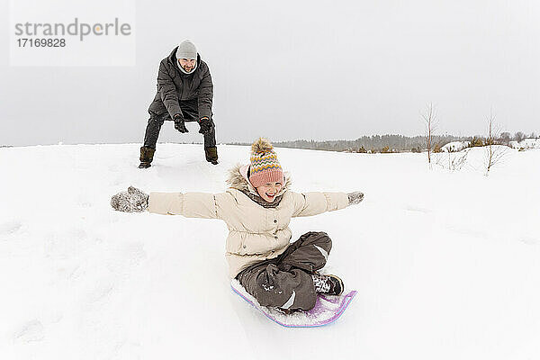 Vater betrachtet fröhliche Tochter beim Schlittenfahren auf verschneitem Land
