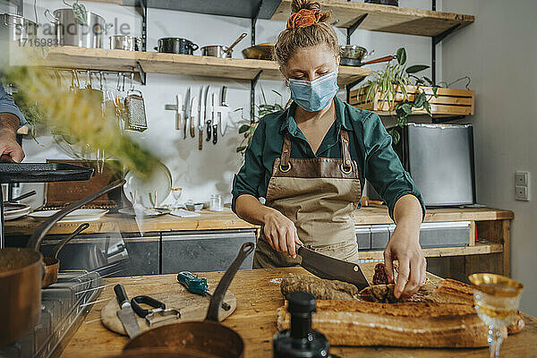 Young chef wearing protective face mask cutting salami while standing in kitchen