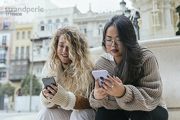 Multi-ethnic female friends using smart phone while sitting against fountain