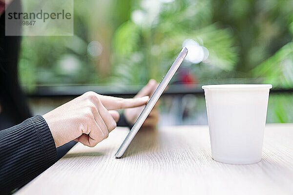 Young woman using digital tablet by coffee cup at table