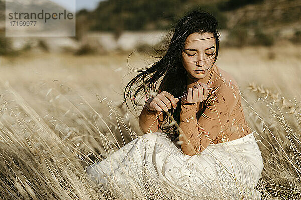 Junge Frau  die in einem windigen Tag inmitten von Gras auf einem Feld sitzt und nachdenkt