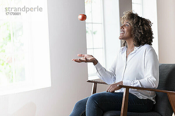 Lächelnde Frau  die mit einem Apfel spielt  während sie zu Hause auf einem Sessel sitzt