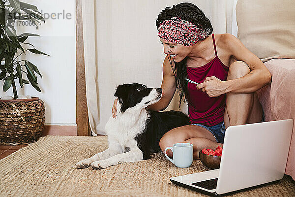 Lächelnde Frau mit Mobiltelefon  die einen Hund betrachtet  während sie zu Hause auf dem Teppich neben einem Laptop sitzt