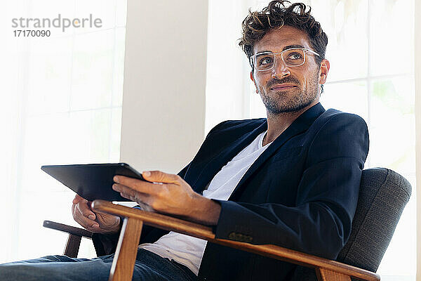Selbstbewusster Geschäftsmann mit digitalem Tablet  der wegschaut  während er zu Hause im Sessel sitzt