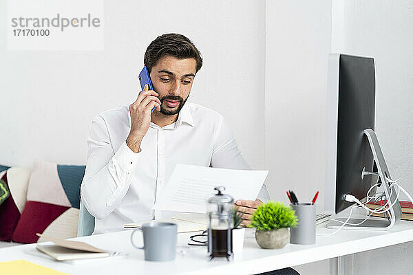 Männlicher Fachmann  der mit einem Smartphone spricht  während er ein Papierdokument im Büro prüft