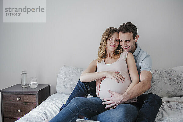 Lächelnder Mann berührt den Bauch seiner schwangeren Frau  während er zu Hause auf dem Bett sitzt