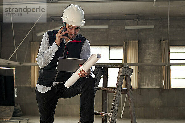 Männlicher Ingenieur  der über ein Mobiltelefon spricht  während er ein digitales Tablet in einem Gebäude benutzt