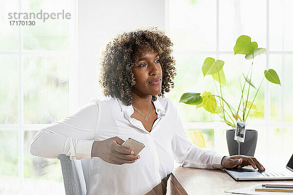 Nachdenkliche Geschäftsfrau mit Laptop und Mobiltelefon  die wegschaut  während sie zu Hause im Büro sitzt
