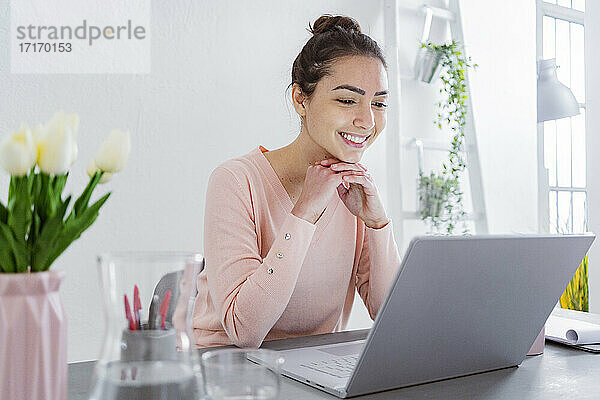 Young woman with hand on chin using laptop while talking with friend on laptop at home