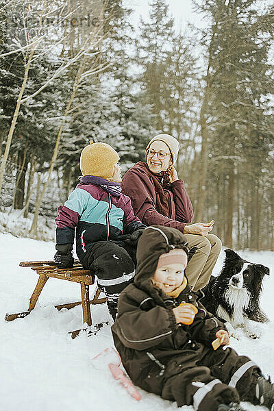 Lächelnde Mutter mit sitzender Tochter bei einem Wochenende mit Familie und Border Collie im Winter im Schnee