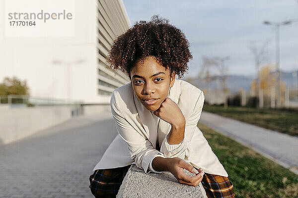 Selbstbewusste junge Frau mit Afro-Haar sitzt auf einer Stützmauer in der Stadt