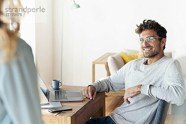 Lächelnder Mann mit Brille  der seine Freundin ansieht  während er zu Hause im Büro sitzt